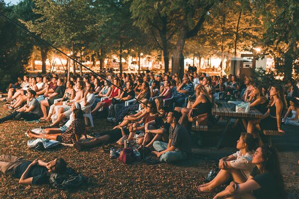 Volxkino/ Altes AKH / Stefan Dvorak/ Echtlicht Menschen sitzen am Abend auf dem Boden im Freien und schauen einen Film auf einer großen Leinwand.