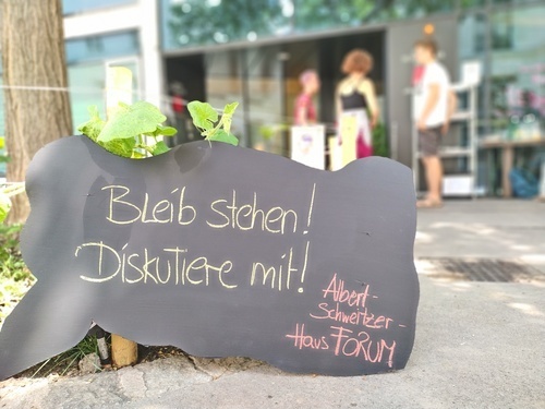 Schild mit der Aufschrift 'Bleib stehen! Diskutiere mit'. Im hintergrund drei menschen, die sich angeregt unterhalten.
