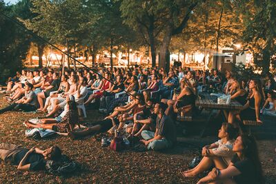 Volxkino/ Altes AKH / Stefan Dvorak/ Echtlicht Menschen sitzen am Abend auf dem Boden im Freien und schauen einen Film auf einer großen Leinwand.